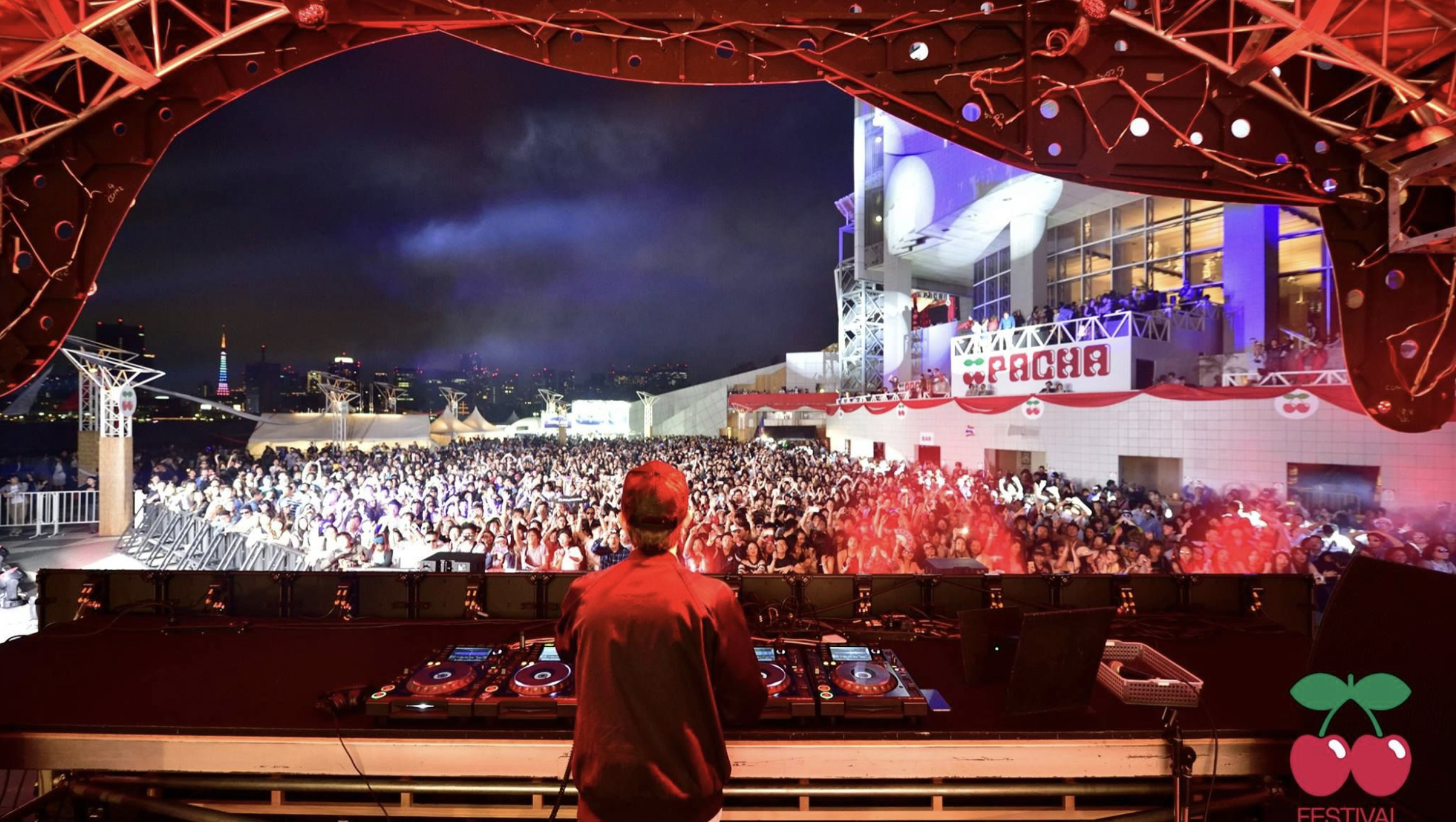 A DJ performs on the Pacha Festival stage overlooking a packed crowd and Tokyo's illuminated waterfront skyline.